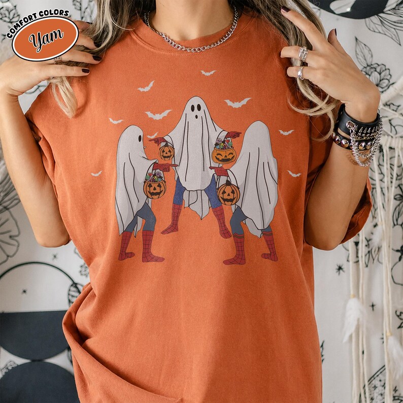 May include: Orange Comfort Colors t-shirt with a Halloween-themed design. The graphic depicts three ghosts dressed as superheroes, carrying pumpkin baskets, with bats overhead. The shirt has a relaxed fit.