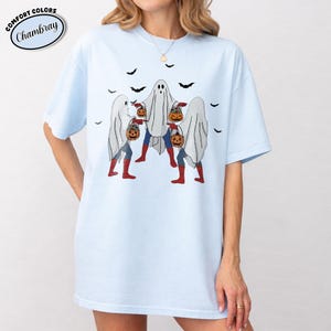 May include: Pale blue t-shirt with a Halloween graphic. The design depicts three figures in ghost costumes, with superhero outfits visible beneath, carrying jack-o'-lanterns. Black bats are scattered above. The shirt has the text "Comfort Colors Chambray" in a circle.