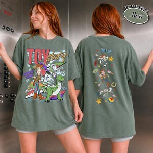 K&ouml;nnte beinhalten: Ein moosgr&uuml;nes T-Shirt mit einer gro&szlig;en Toy Story-Grafik auf der Vorderseite, die Woody, Buzz Lightyear und Rex zeigt, sowie dem Text "Toy Story Since 1995". Die R&uuml;ckseite zeigt eine vertikale Anordnung kleinerer Charakter-Icons und Sterne.