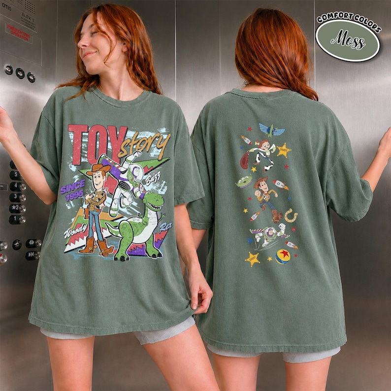 K&ouml;nnte beinhalten: Ein moosgr&uuml;nes T-Shirt mit einer gro&szlig;en Toy Story-Grafik auf der Vorderseite, die Woody, Buzz Lightyear und Rex zeigt, sowie dem Text "Toy Story Since 1995". Die R&uuml;ckseite zeigt eine vertikale Anordnung kleinerer Charakter-Icons und Sterne.