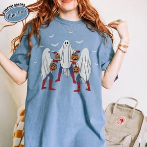 May include: Pale blue t-shirt with a Halloween design. The graphic shows three figures in ghost costumes, carrying pumpkin-shaped lanterns. They wear red boots and blue trousers. The shirt has the words "Comfort Colors" and "Blue Jean" in a circle.