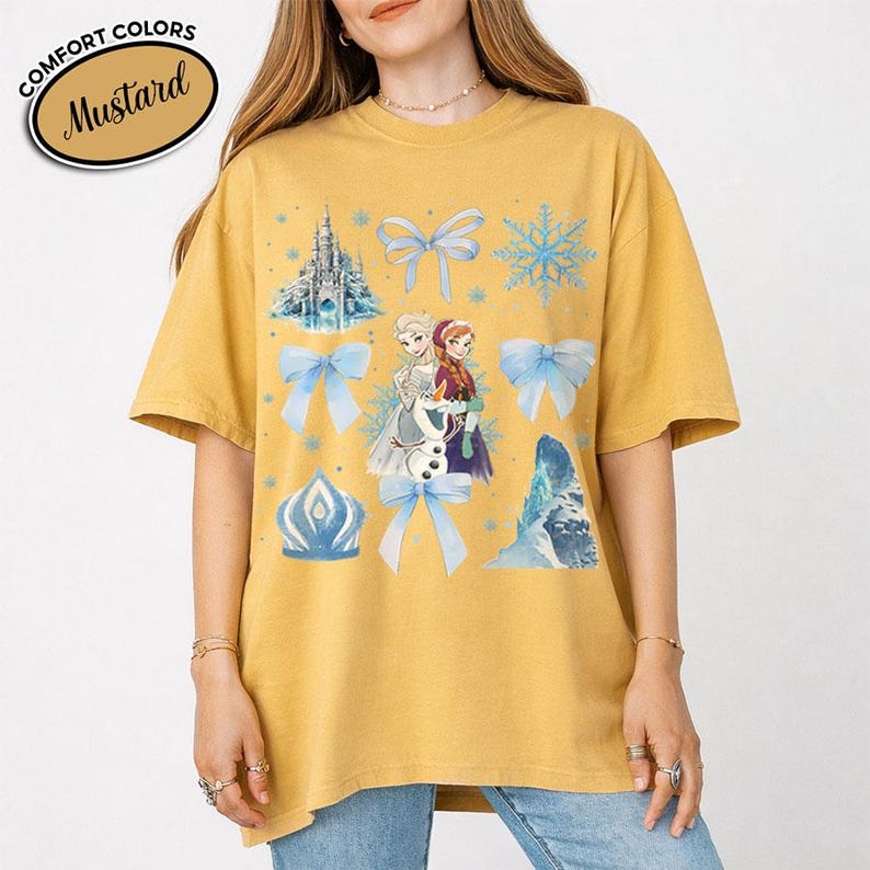 Puede incluir: Camiseta de color mostaza con un gr&aacute;fico de dos personajes femeninos animados, un mu&ntilde;eco de nieve, un castillo, copos de nieve y lazos. La camiseta tiene las palabras "COMFORT COLORS" y "Mustard" en un dise&ntilde;o circular.