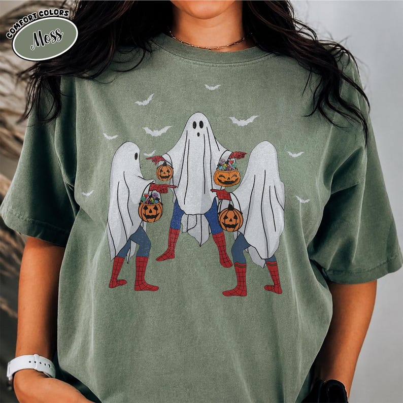 May include: A moss green t-shirt with a Halloween-themed graphic. The design depicts three ghosts in superhero costumes, carrying pumpkin-shaped candy buckets. Bats are in the background. The text "COMFORT COLORS" and "Moss" is in the upper left corner.
