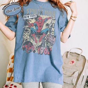 Puede incluir: Camiseta azul con un gr&aacute;fico vintage de Spider-Man. El dise&ntilde;o incluye el texto "The Amazing Spider-Man", "New York" y "Swing Into Action 1962". La camiseta tiene una est&eacute;tica retro y desgastada.