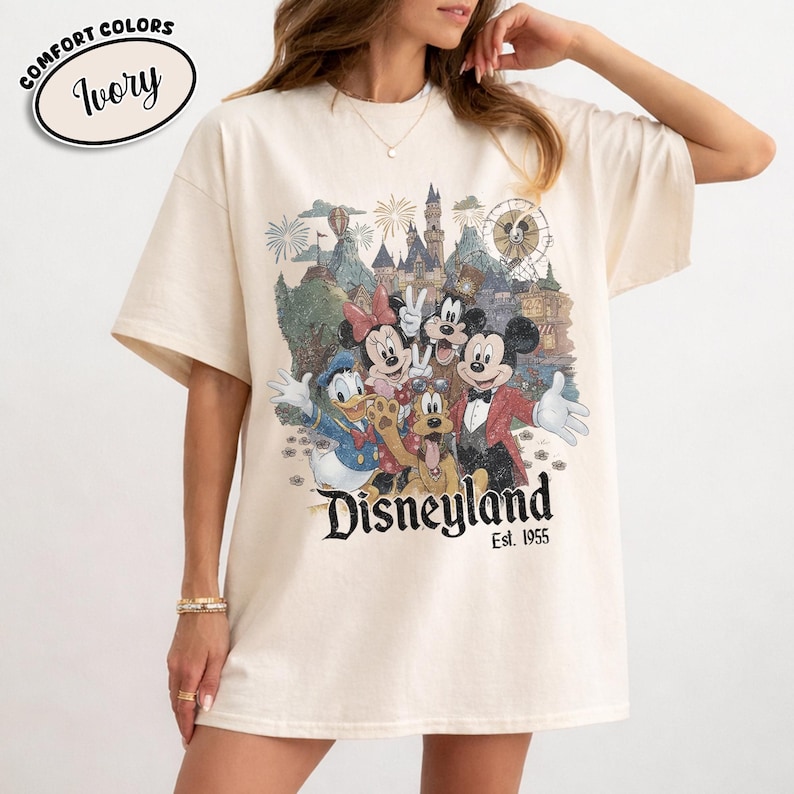 Może przedstawiać: Kremowa koszulka z grafiką Disneyland z Myszką Miki, Minnie, Kaczorem Donaldem, Goofym i Pluto. Grafika zawiera zamek, fajerwerki i diabelski młyn. Tekst "Disneyland Est. 1955" jest wydrukowany poniżej.