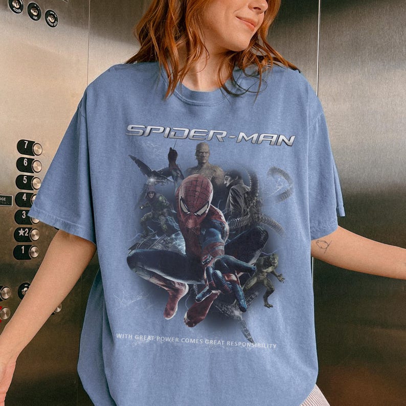 May include: Pale blue Spider-Man t-shirt with a graphic of Spider-Man and other characters. The text "SPIDER-MAN" is across the top, and "WITH GREAT POWER COMES GREAT RESPONSIBILITY" is at the bottom.