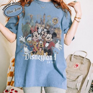 Może przedstawiać: Niebieska koszulka w kolorze dżinsu z grafiką Disneyland z Myszką Miki, Minnie, Kaczorem Donaldem, Goofym i innymi postaciami. Grafika zawiera zamek, diabelski młyn i napis "Disneyland Est. 1955."
