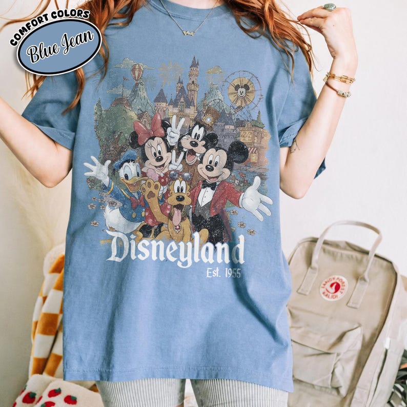 Może przedstawiać: Niebieska koszulka w kolorze dżinsu z grafiką Disneyland z Myszką Miki, Minnie, Kaczorem Donaldem, Goofym i innymi postaciami. Grafika zawiera zamek, diabelski młyn i napis "Disneyland Est. 1955."
