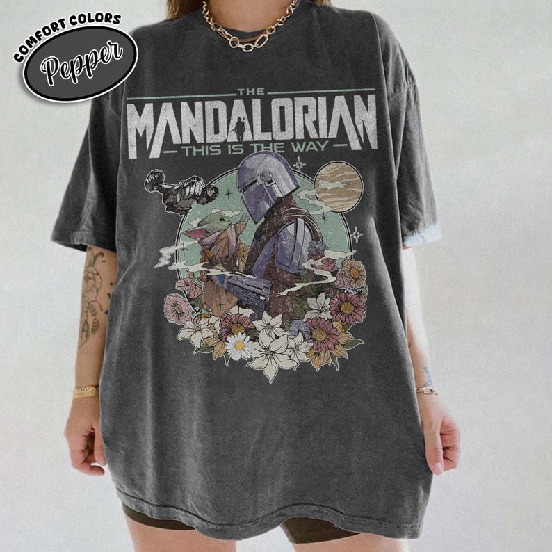 May include: Dark gray t-shirt featuring a graphic of The Mandalorian and Grogu surrounded by flowers. The text "THE MANDALORIAN - THIS IS THE WAY" is printed above the image. The shirt has a Comfort Colors Pepper label.
