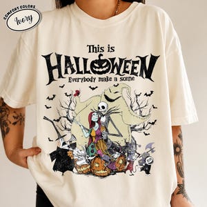 Puede incluir: Camiseta de color marfil con un dise&ntilde;o tem&aacute;tico de Halloween. El dise&ntilde;o incluye el texto "This is Halloween" con una calabaza, murci&eacute;lagos y personajes de la pel&iacute;cula. La camiseta tiene cuello redondo y mangas cortas.