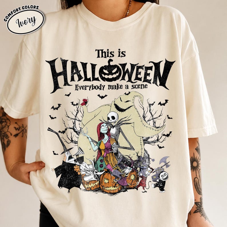 Puede incluir: Camiseta de color marfil con un dise&ntilde;o tem&aacute;tico de Halloween. El dise&ntilde;o incluye el texto "This is Halloween" con una calabaza, murci&eacute;lagos y personajes de la pel&iacute;cula. La camiseta tiene cuello redondo y mangas cortas.