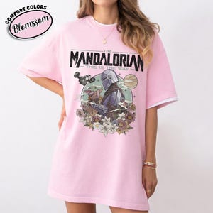 May include: Pink t-shirt featuring a graphic of The Mandalorian and Grogu surrounded by flowers. The text "THE MANDALORIAN" and "THIS IS THE WAY" are printed above the image. The shirt has white trim on the sleeves.