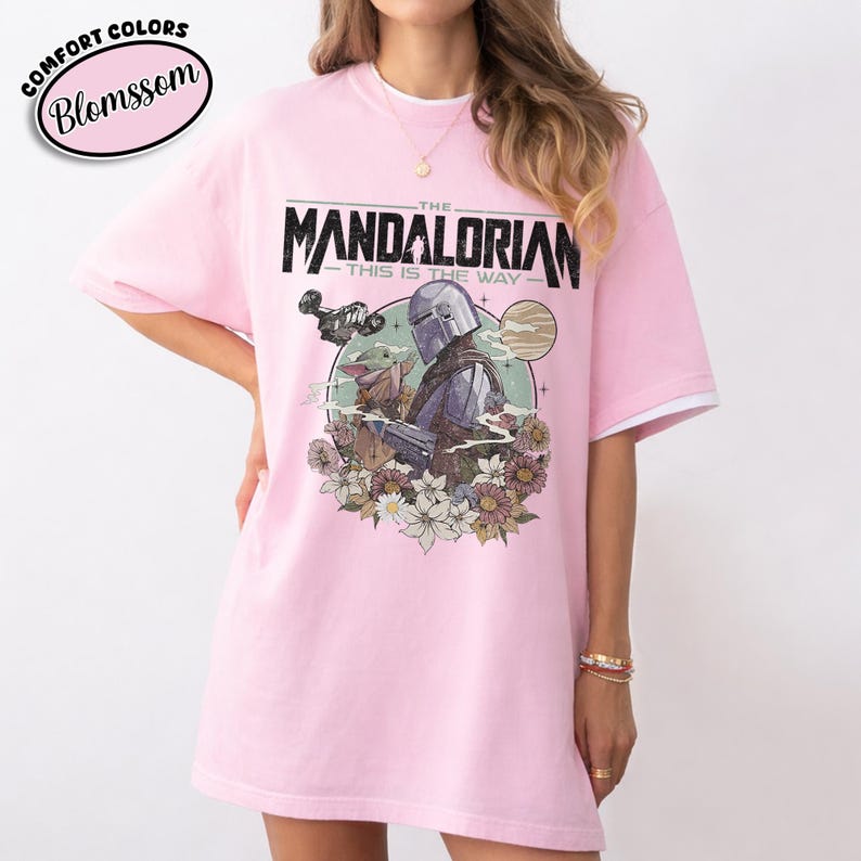 May include: Pink t-shirt featuring a graphic of The Mandalorian and Grogu surrounded by flowers. The text "THE MANDALORIAN" and "THIS IS THE WAY" are printed above the image. The shirt has white trim on the sleeves.