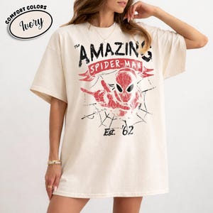May include: Ivory-colored t-shirt featuring a distressed graphic of Spider-Man with the text "The Amazing Spider-Man" and "Est. '62". The shirt has a crew neck and short sleeves. The words "Comfort Colors" and "Ivory" are in a circle at the top left.