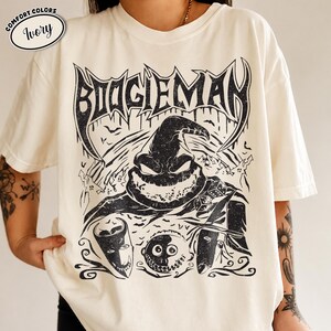 K&ouml;nnte beinhalten: Elfenbeinfarbenes T-Shirt mit einer schwarzen Grafik der Figur Boogieman. Das Design enth&auml;lt das Wort "BOOGIEMAN" in einer stilisierten Schriftart sowie Flederm&auml;use, W&uuml;rfel und andere Bilder. Das Shirt ist im Comfort-Colors-Stil.