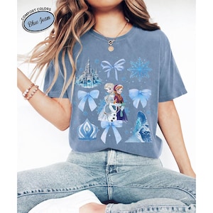 Puede incluir: Camiseta azul vaquero con un gr&aacute;fico de los personajes Elsa y Anna de la pel&iacute;cula Frozen, junto con copos de nieve, lazos y un castillo. La camiseta tiene el texto "Comfort Colors Blue Jean" en un dise&ntilde;o circular.