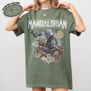 May include: Moss green t-shirt featuring a graphic of The Mandalorian and Grogu surrounded by flowers. The text "THE MANDALORIAN - THIS IS THE WAY" is printed across the front. The shirt has a relaxed fit.