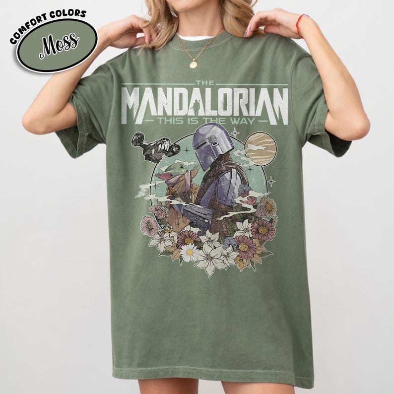 May include: Moss green t-shirt featuring a graphic of The Mandalorian and Grogu surrounded by flowers. The text "THE MANDALORIAN - THIS IS THE WAY" is printed across the front. The shirt has a relaxed fit.