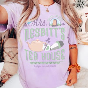 K&ouml;nnte beinhalten: Ein helllila T-Shirt mit dem Text "Mrs. Nesbitt's Tea House" in Gr&uuml;n und Wei&szlig;. Das Design umfasst eine Teekanne, eine Figur in einer Blase und den Satz "To Infini-tea and Beyond."