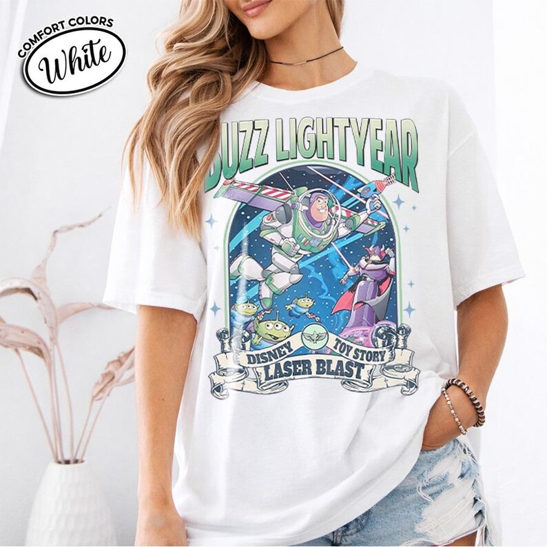 May include: White Comfort Colors t-shirt featuring a graphic of Buzz Lightyear from Toy Story. The design includes Buzz in a spaceship, aliens, and the text "Buzz Lightyear" and "Laser Blast".