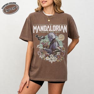 May include: Espresso-colored t-shirt featuring a graphic of The Mandalorian with Grogu, surrounded by flowers. The text on the shirt reads "THE MANDALORIAN - THIS IS THE WAY". The shirt has a vintage, worn look.