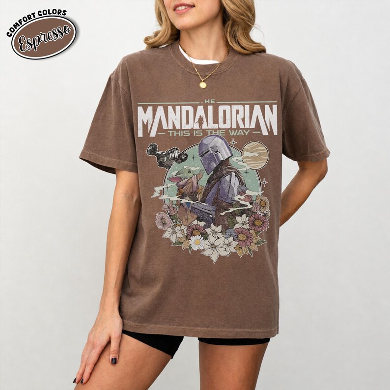 May include: Espresso-colored t-shirt featuring a graphic of The Mandalorian with Grogu, surrounded by flowers. The text on the shirt reads "THE MANDALORIAN - THIS IS THE WAY". The shirt has a vintage, worn look.