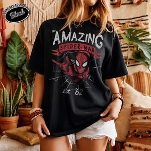 May include: Black t-shirt featuring a distressed graphic of Spider-Man with the text "THE AMAZING SPIDER-MAN" and "Est. '62". The shirt has a vintage look and feel, with a red and white color scheme. The words "COMFORT COLORS" and "Black" are printed on the top left.