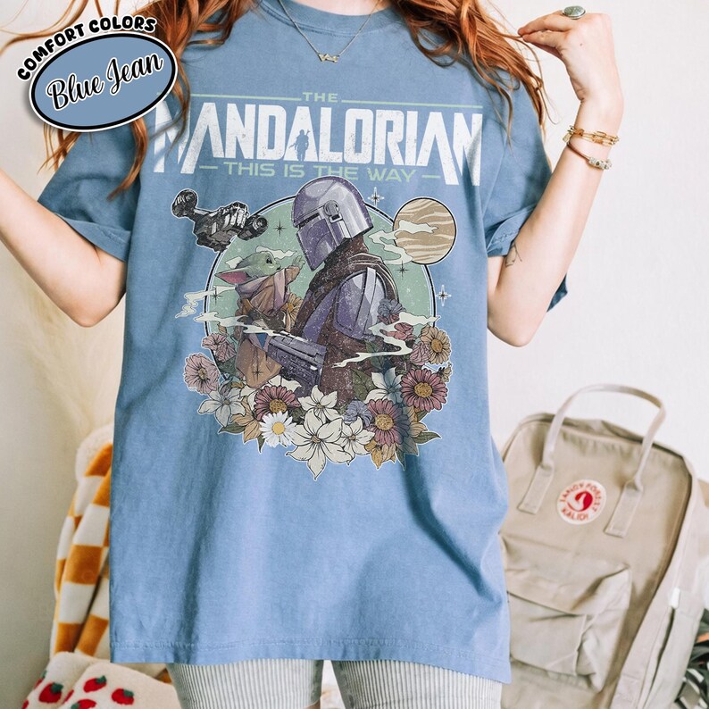 May include: Blue t-shirt featuring a graphic of The Mandalorian and Grogu surrounded by flowers. The text "THE MANDALORIAN" and "THIS IS THE WAY" are printed above the image. The shirt has short sleeves and a crew neck.