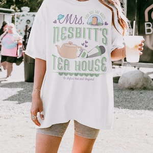K&ouml;nnte beinhalten: Wei&szlig;es T-Shirt mit einem Grafikdesign f&uuml;r "Mrs. Nesbitt's Tea House". Das Design umfasst eine Teekanne, einen Hut und den Text "To Infini-tea and Beyond". Das Shirt ist &uuml;bergro&szlig; und hat kurze &Auml;rmel.