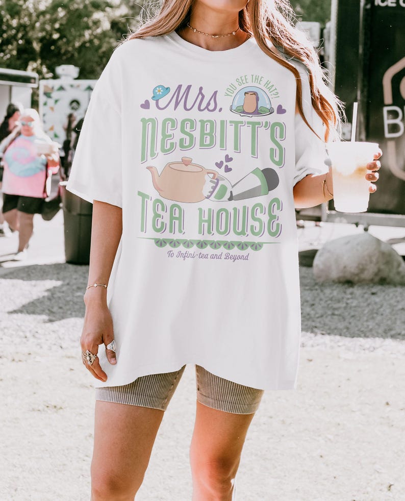 K&ouml;nnte beinhalten: Wei&szlig;es T-Shirt mit einem Grafikdesign f&uuml;r "Mrs. Nesbitt's Tea House". Das Design umfasst eine Teekanne, einen Hut und den Text "To Infini-tea and Beyond". Das Shirt ist &uuml;bergro&szlig; und hat kurze &Auml;rmel.