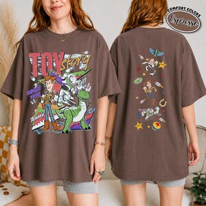K&ouml;nnte beinhalten: Ein espresso-braunes T-Shirt mit einer gro&szlig;en Toy Story-Grafik auf der Vorderseite, die Woody, Buzz Lightyear und Rex sowie den Text Toy Story Since 1995 zeigt. Die R&uuml;ckseite zeigt eine vertikale Anordnung kleinerer Charakterillustrationen.