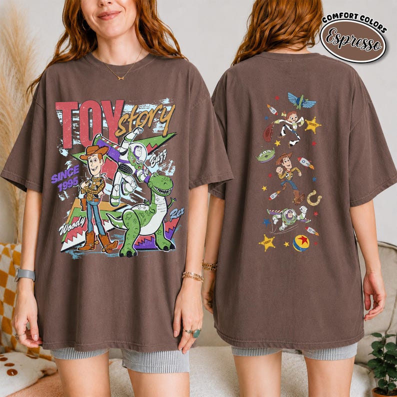 K&ouml;nnte beinhalten: Ein espresso-braunes T-Shirt mit einer gro&szlig;en Toy Story-Grafik auf der Vorderseite, die Woody, Buzz Lightyear und Rex sowie den Text Toy Story Since 1995 zeigt. Die R&uuml;ckseite zeigt eine vertikale Anordnung kleinerer Charakterillustrationen.