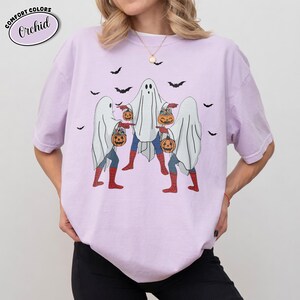 May include: A lilac t-shirt featuring a graphic of three ghosts dressed as superheroes, each carrying a pumpkin. The shirt has the text "COMFORT COLORS" and "Orchid" in a circle. Black bats are scattered around the ghosts.