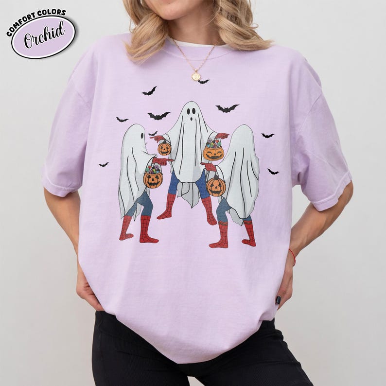 May include: A lilac t-shirt featuring a graphic of three ghosts dressed as superheroes, each carrying a pumpkin. The shirt has the text "COMFORT COLORS" and "Orchid" in a circle. Black bats are scattered around the ghosts.