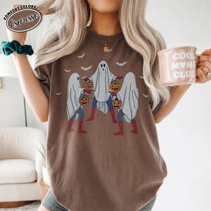 May include: Espresso-coloured t-shirt with a Halloween graphic. The design shows three ghosts holding jack-o'-lanterns, with bat silhouettes above. The shirt has a relaxed fit and is made of a soft material.