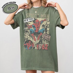 Puede incluir: Camiseta verde oliva con un gr&aacute;fico vintage de Spider-Man. El dise&ntilde;o incluye las palabras "The Amazing Spider-Man", "New York" y "1962". El gr&aacute;fico representa a Spider-Man en acci&oacute;n con un estilo retro de c&oacute;mic.