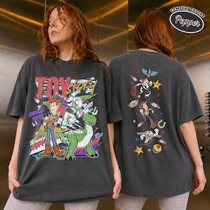 K&ouml;nnte beinhalten: Ein anthrazitfarbenes T-Shirt mit einer Grafik auf der Vorderseite von Woody, Buzz Lightyear und Rex sowie dem Text Toy Story Since 1995. Die R&uuml;ckseite zeigt eine vertikale Anordnung verschiedener Figuren und Symbole aus dem Film, darunter Woody und Buzz.