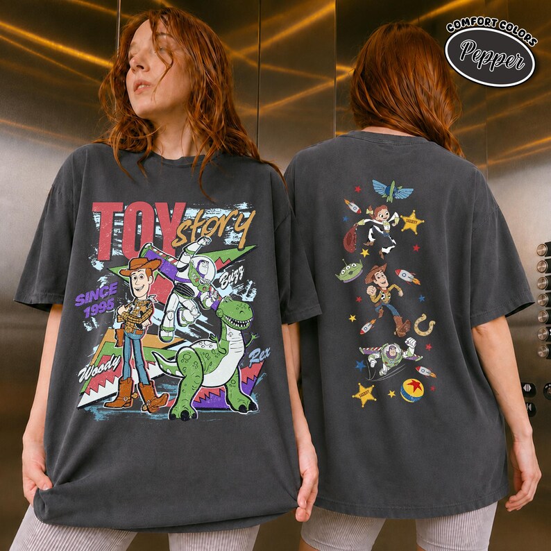 K&ouml;nnte beinhalten: Ein anthrazitfarbenes T-Shirt mit einer Grafik auf der Vorderseite von Woody, Buzz Lightyear und Rex sowie dem Text Toy Story Since 1995. Die R&uuml;ckseite zeigt eine vertikale Anordnung verschiedener Figuren und Symbole aus dem Film, darunter Woody und Buzz.