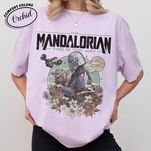 May include: Orchid-colored t-shirt featuring a graphic of The Mandalorian and Grogu, with the text "THE MANDALORIAN - THIS IS THE WAY." The design includes a spaceship, planet, and floral elements. The shirt has short sleeves and a crew neck.