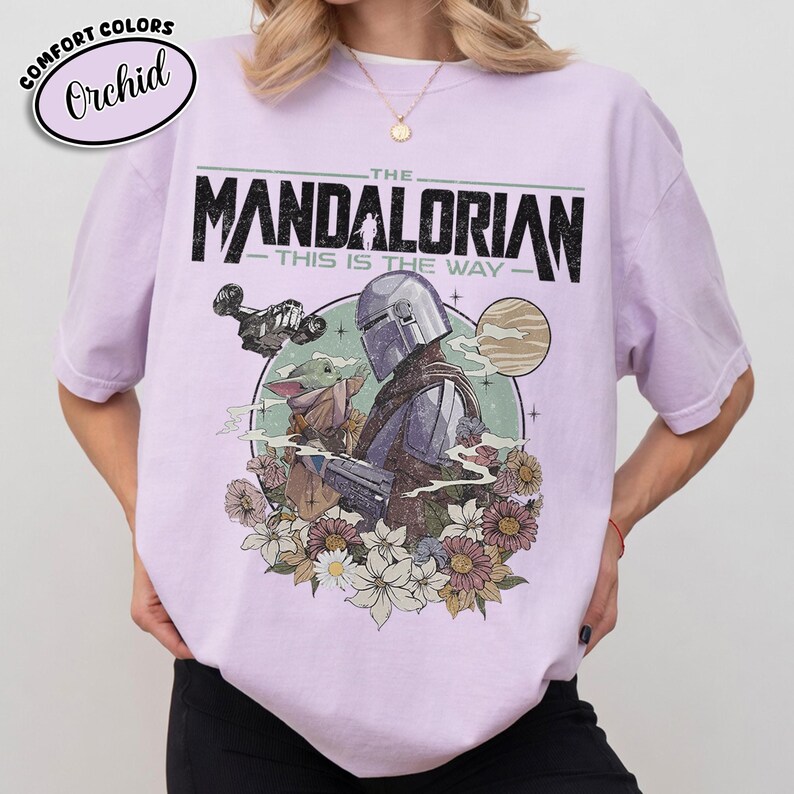 May include: Orchid-colored t-shirt featuring a graphic of The Mandalorian and Grogu, with the text "THE MANDALORIAN - THIS IS THE WAY." The design includes a spaceship, planet, and floral elements. The shirt has short sleeves and a crew neck.