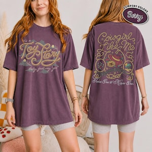 K&ouml;nnte beinhalten: Ein beerenfarbenes Kurzarm-T-Shirt mit einer Vorderseiten-Grafik, die den Text Toy Story in einer seilartigen Schrift zeigt. Die R&uuml;ckseite zeigt den Text Cowgirl Like Me und Takes One to Know One, umgeben von Seilen, einem Hut und W&uuml;stenmotiven.