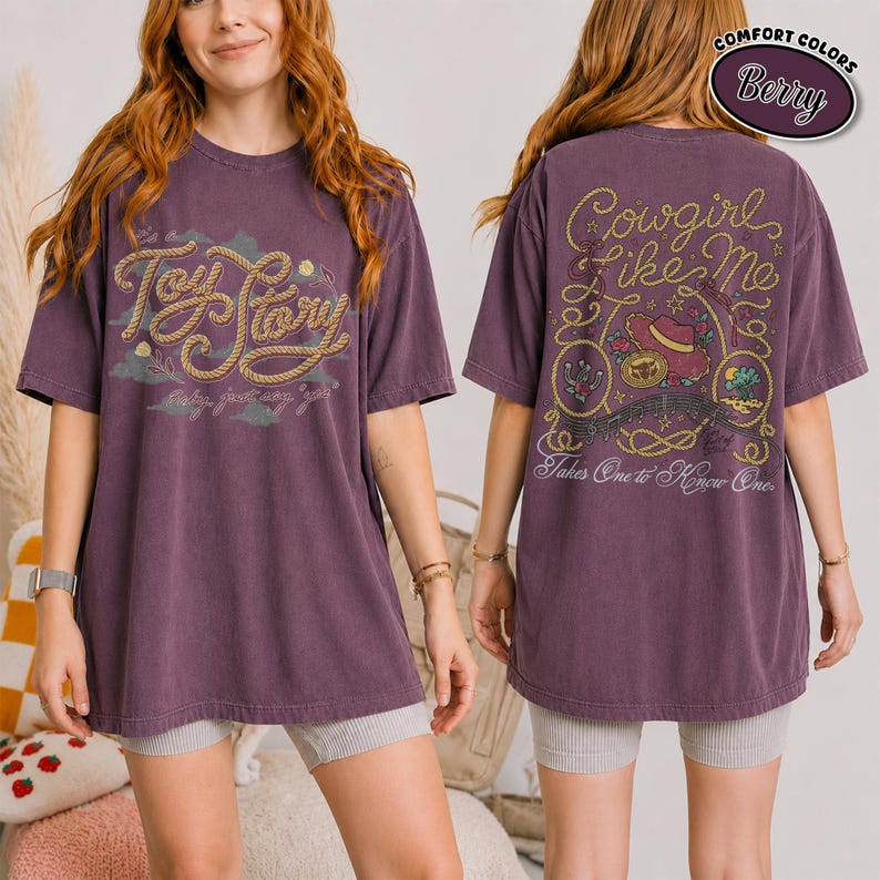 K&ouml;nnte beinhalten: Ein beerenfarbenes Kurzarm-T-Shirt mit einer Vorderseiten-Grafik, die den Text Toy Story in einer seilartigen Schrift zeigt. Die R&uuml;ckseite zeigt den Text Cowgirl Like Me und Takes One to Know One, umgeben von Seilen, einem Hut und W&uuml;stenmotiven.