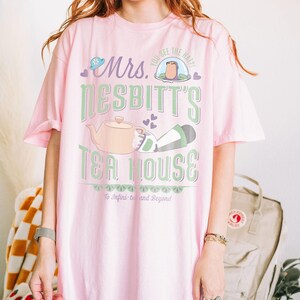 K&ouml;nnte beinhalten: Hellrosa T-Shirt mit dem Text "Mrs. Nesbitt's Tea House" in Gr&uuml;n und Wei&szlig;. Das Design beinhaltet eine Teekanne, einen Hut und den Satz "To Infini-tea and Beyond".