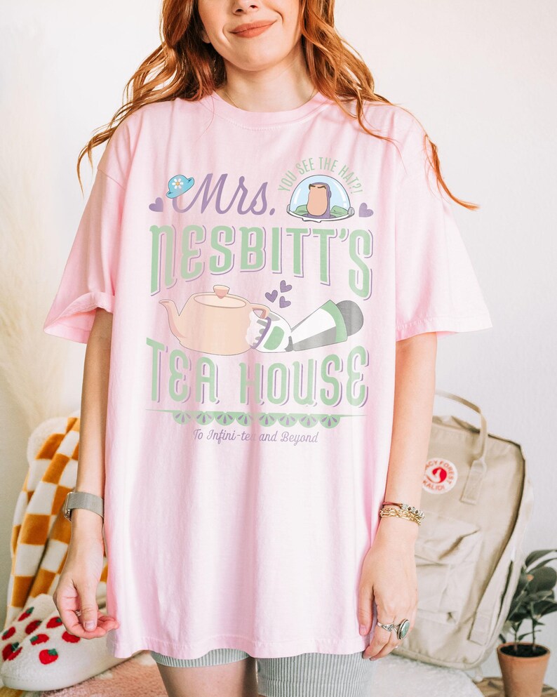 K&ouml;nnte beinhalten: Hellrosa T-Shirt mit dem Text "Mrs. Nesbitt's Tea House" in Gr&uuml;n und Wei&szlig;. Das Design beinhaltet eine Teekanne, einen Hut und den Satz "To Infini-tea and Beyond".