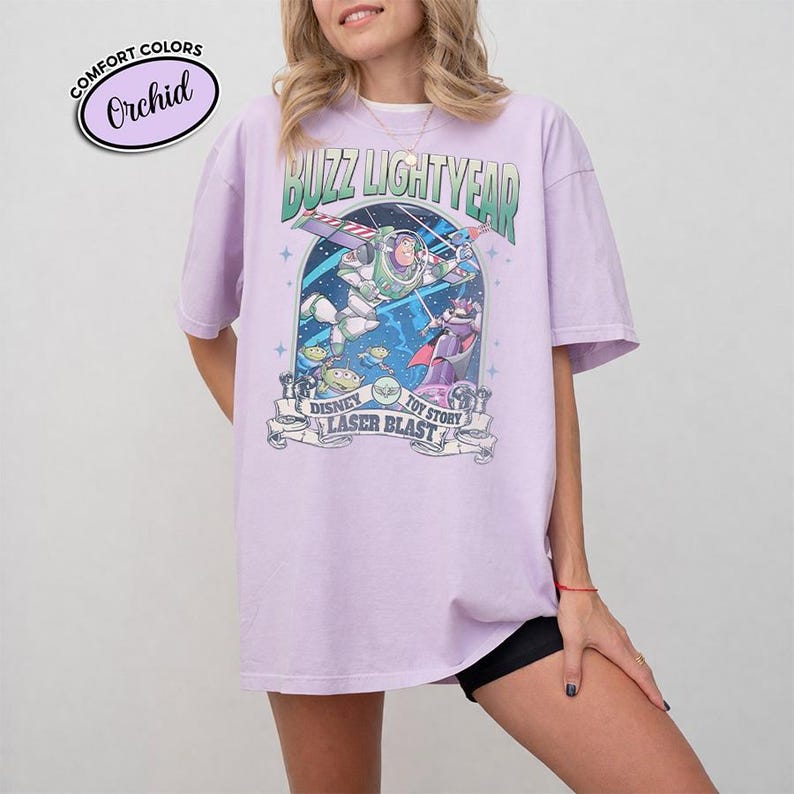 May include: A light purple t-shirt featuring a graphic of Buzz Lightyear from Disney's Toy Story. The design includes the text "Buzz Lightyear", "Disney Toy Story", and "Laser Blast". The shirt is a Comfort Colors brand.
