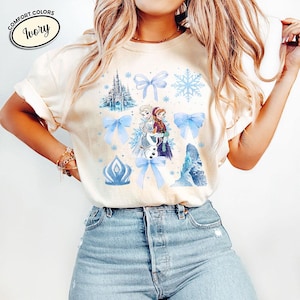 Puede incluir: Camiseta de color marfil con un gr&aacute;fico de personajes de dibujos animados, lazos azules, copos de nieve y un castillo. La camiseta tiene las palabras "Comfort Colors" e "Ivory" impresas.