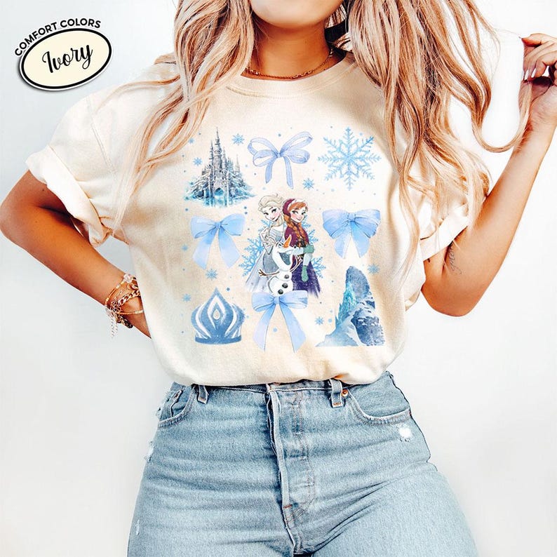 Puede incluir: Camiseta de color marfil con un gr&aacute;fico de personajes de dibujos animados, lazos azules, copos de nieve y un castillo. La camiseta tiene las palabras "Comfort Colors" e "Ivory" impresas.
