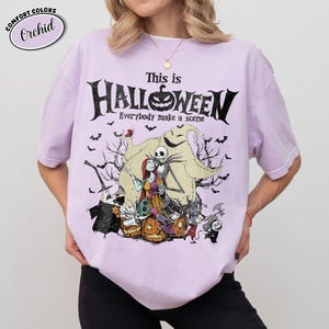 Puede incluir: Una camiseta de color lila claro con un gr&aacute;fico de "Pesadilla antes de Navidad". El dise&ntilde;o presenta a Jack Skellington, Sally y otros personajes con el texto "This is Halloween". La camiseta es de la marca Comfort Colors.