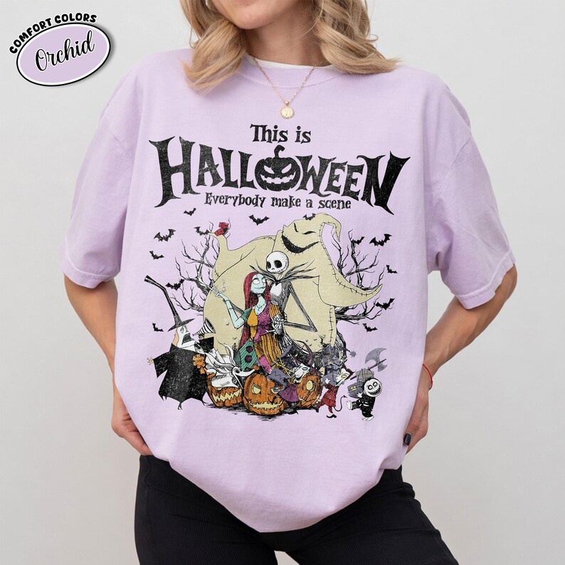 Puede incluir: Una camiseta de color lila claro con un gr&aacute;fico de "Pesadilla antes de Navidad". El dise&ntilde;o presenta a Jack Skellington, Sally y otros personajes con el texto "This is Halloween". La camiseta es de la marca Comfort Colors.