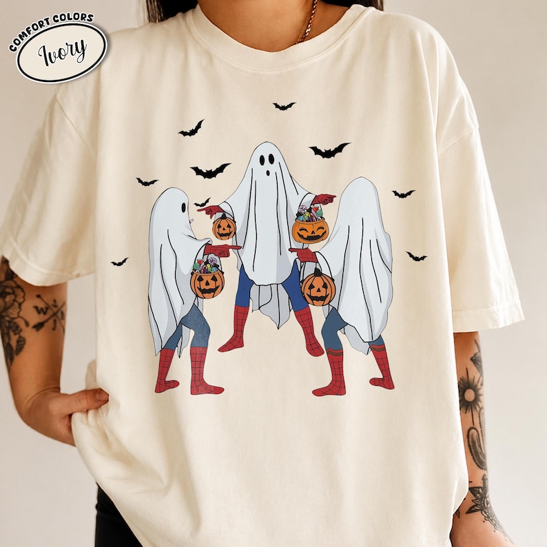 May include: Ivory-colored t-shirt featuring a Halloween-themed design. Three figures dressed as ghosts are holding jack-o'-lantern buckets. The figures have Spider-Man-inspired legs and are surrounded by black bat silhouettes. The shirt has the words "Comfort Colors" and "Ivory" in a circle.